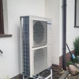 Zewnętrzna jednostka pompy ciepła Panasonic zamontowana na białej stalowej konstrukcji, na betonowych fundamentach przy ścianie domu z brązowymi elementami dekoracyjnymi.