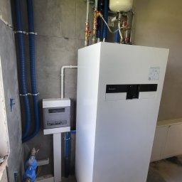 MIKA Przemysław Kowal - Biała pompa ciepła Panasonic, widoczne niebieskie rury instalacyjne, szara płytka na ścianie, skrzynka elektryczna i elementy instalacji hydraulicznej w surowym stanie wykończenia.