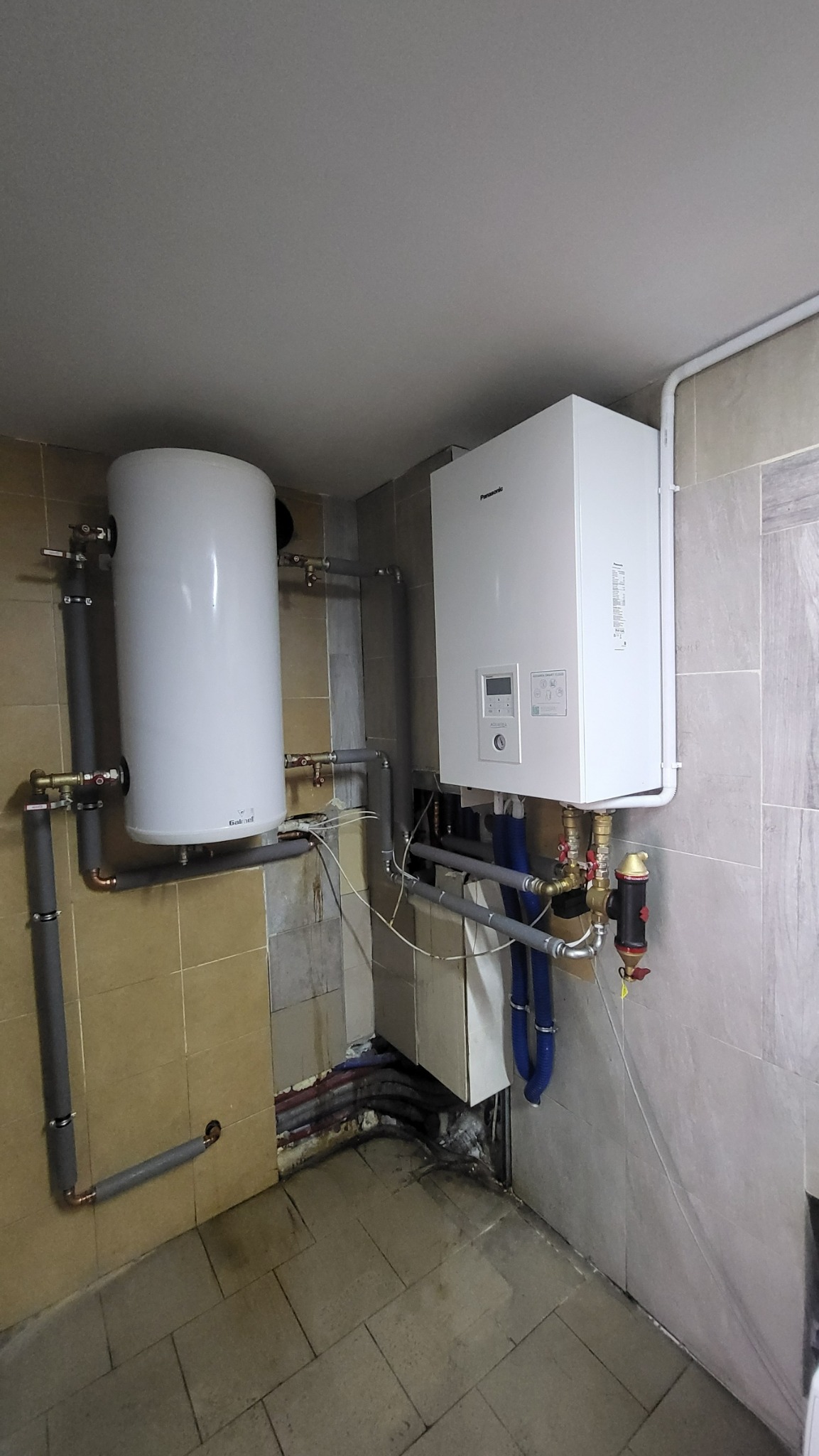 Biały, wiszący piec gazowy marki Panasonic, podłączony do instalacji grzewczej z rurami w izolacji, obok pionowy, biały zasobnik ciepłej wody Galmet, zamontowane w pomieszczeniu z ceramicznymi...