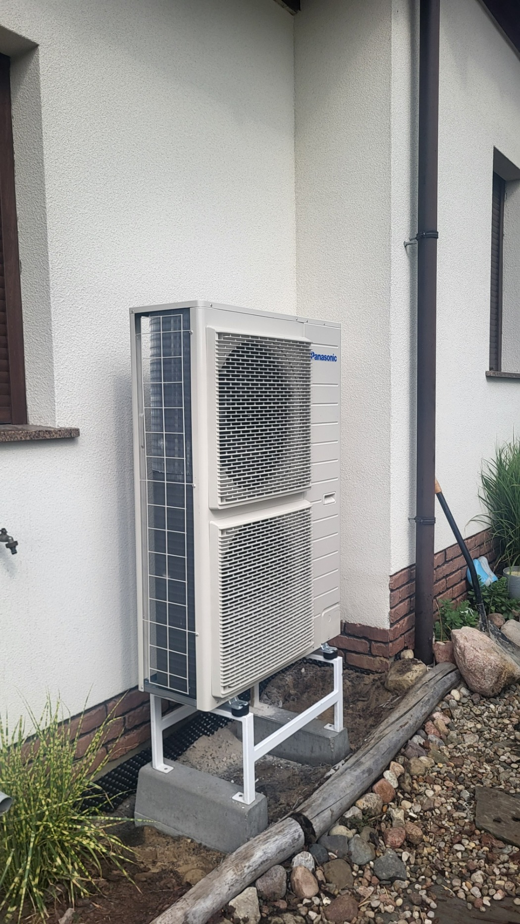 Zewnętrzna jednostka pompy ciepła Panasonic zamontowana na białej stalowej konstrukcji, na betonowych fundamentach przy ścianie domu z brązowymi elementami dekoracyjnymi.