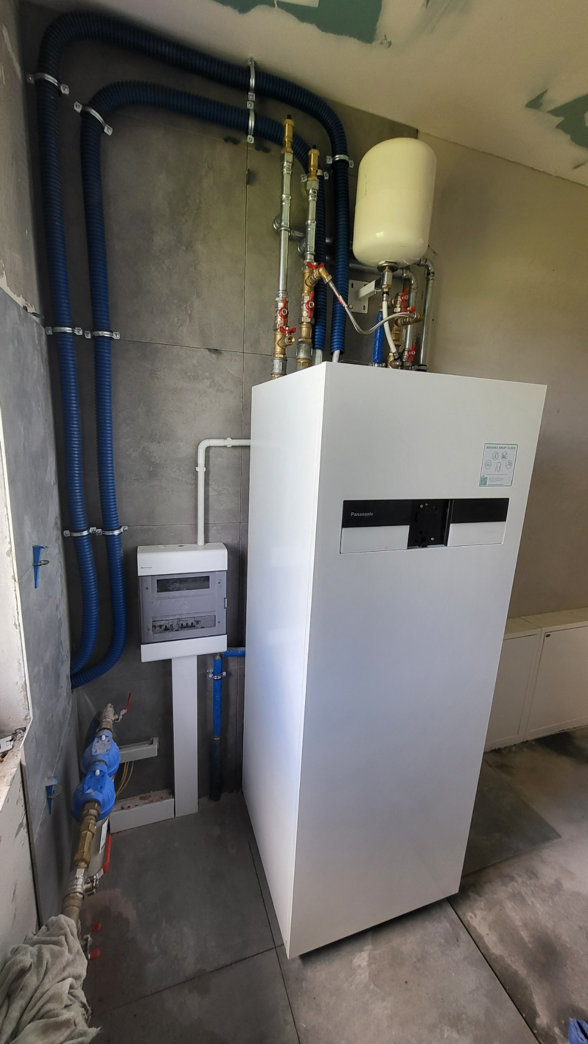 Biała pompa ciepła Panasonic, widoczne niebieskie rury instalacyjne, szara płytka na ścianie, skrzynka elektryczna i elementy instalacji hydraulicznej w surowym stanie wykończenia.