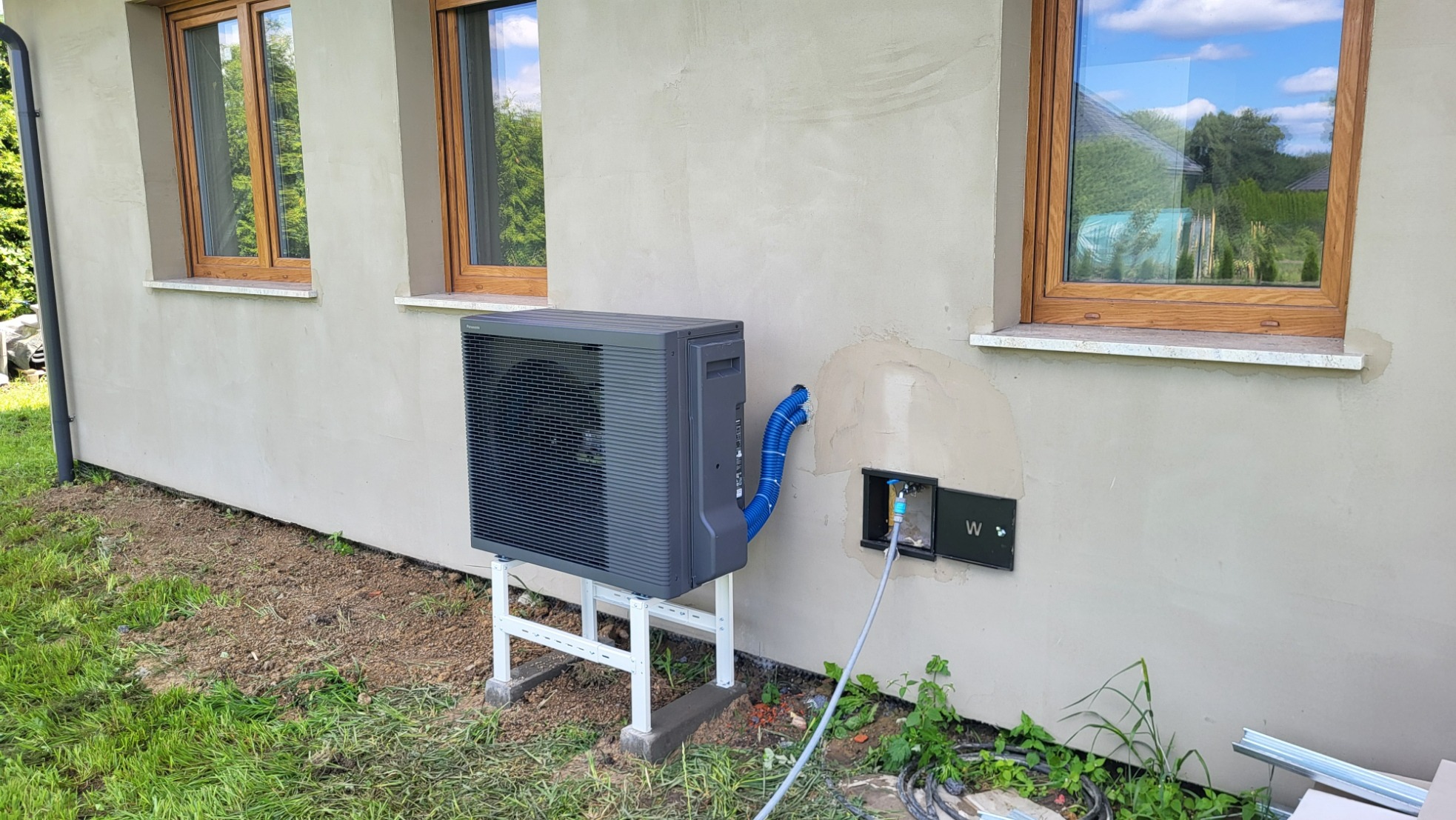 Szara pompa ciepła powietrze-woda zamontowana na białym stelażu przy ścianie budynku z dwoma oknami, widoczne niebieskie rury łączące pompę z instalacją wewnątrz budynku.