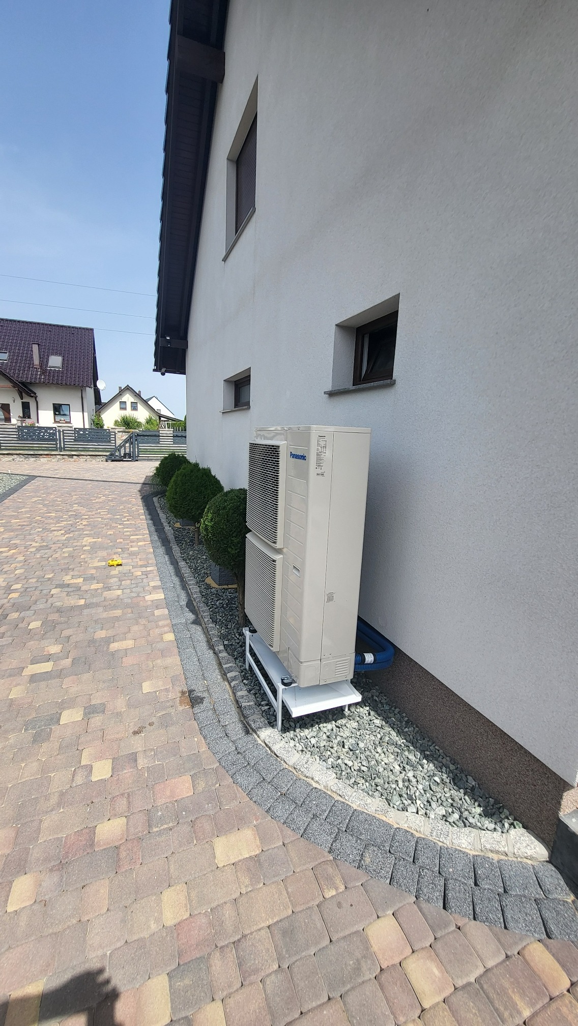 Zewnętrzna jednostka pompy ciepła Panasonic zamontowana na białym stelażu przy ścianie domu z ozdobnym obsypaniem kamieniami i niebieskimi rurami odprowadzającymi.