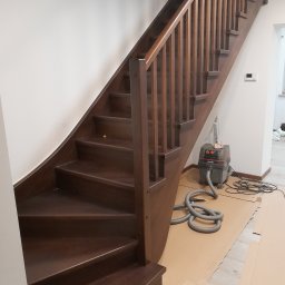 P.P.H.U.Heban - Ciemnobrązowe schody drewniane z balustradą, widoczne zabezpieczenia podłogi kartonem i odkurzacz przemysłowy, sugerujące prace wykończeniowe.