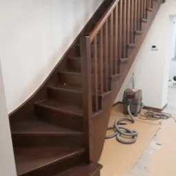 Ciemnobrązowe schody drewniane z balustradą, widoczne zabezpieczenia podłogi kartonem i odkurzacz przemysłowy, sugerujące prace wykończeniowe.
