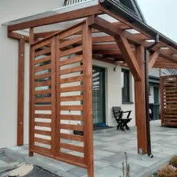 Zadaszenie tarasu z drewnianą konstrukcją i ażurowymi ściankami bocznymi, widoczne rynny, szara kostka brukowa i fragment domu.