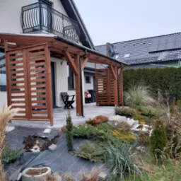 Drewniana konstrukcja z poziomymi listwami tworząca zadaszony taras przy domu jednorodzinnym, widok na ogród z roślinami ozdobnymi i balkon z metalową balustradą.