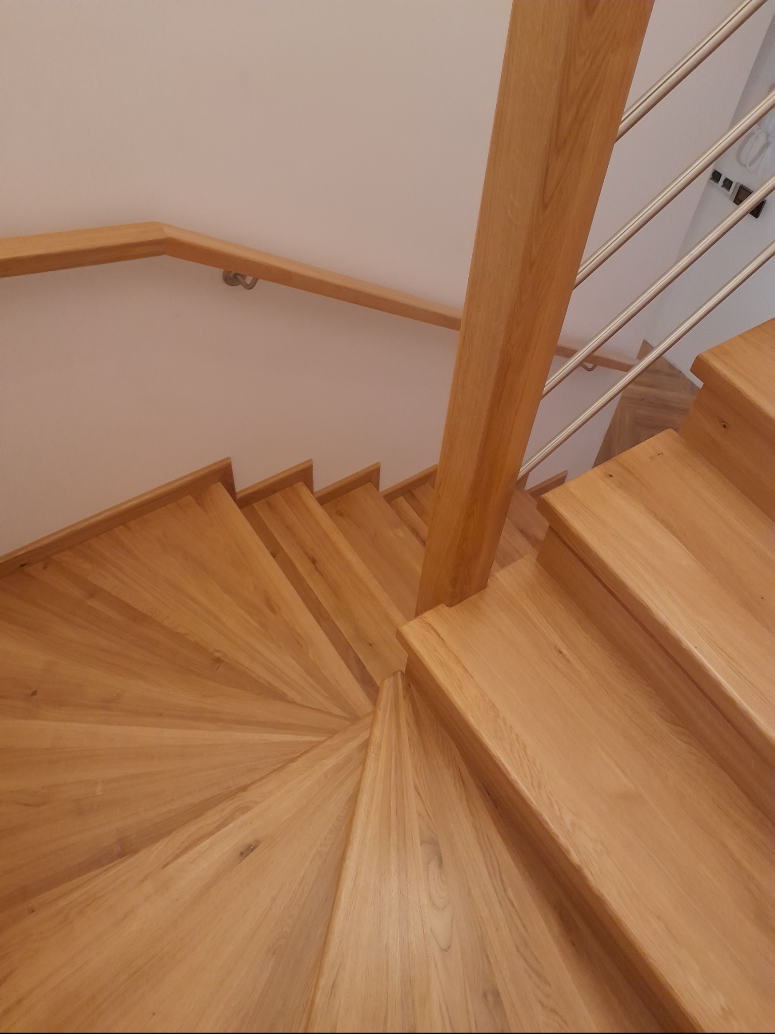 Widok z góry na spiralne, jasne stopnie z drewna z balustradą z poziomych, metalowych prętów i drewnianą poręczą przy ścianie.