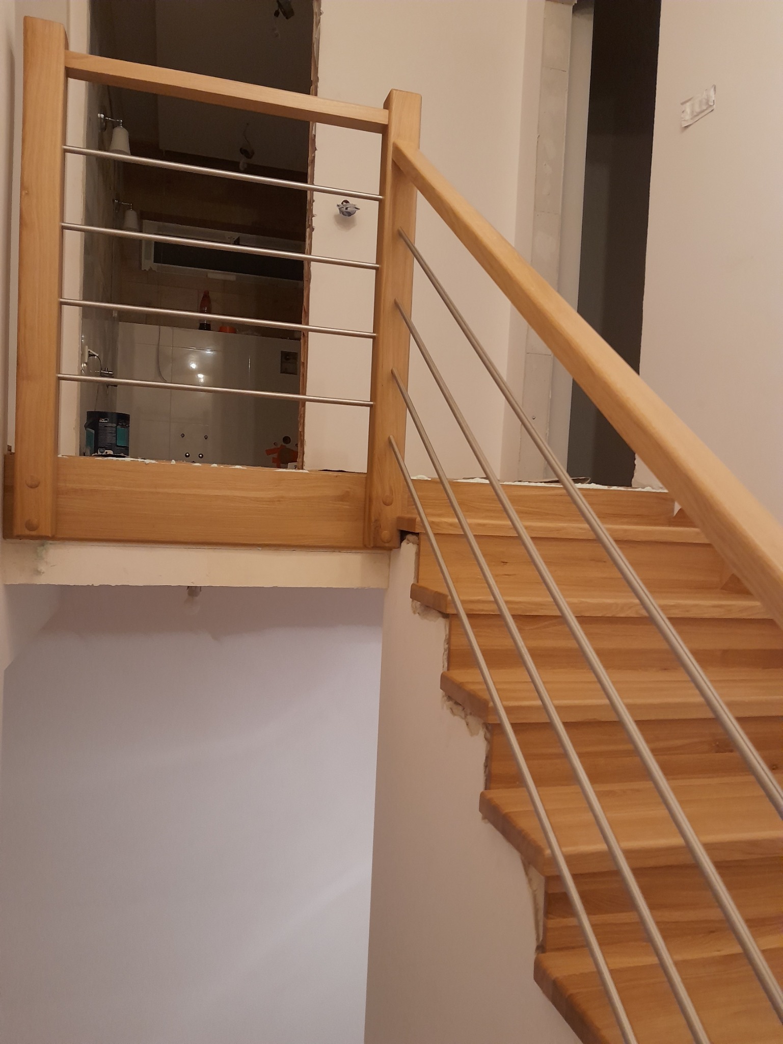 Wnętrze domu w trakcie remontu z widocznymi drewnianymi schodami z metalowymi balustradami i otwartym przejściem do łazienki.