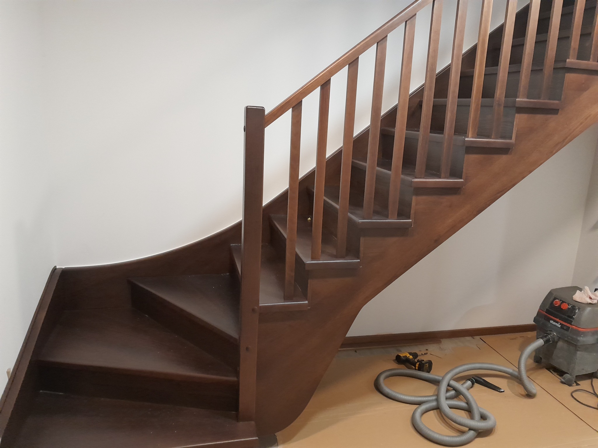 Wykonane z ciemnego drewna schody z balustradą, z zakrętem u dołu, w trakcie prac wykończeniowych, widoczny odkurzacz przemysłowy i wkrętarka.