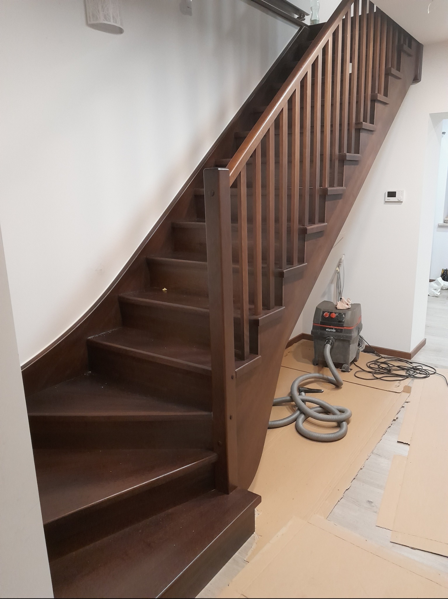 Ciemnobrązowe schody drewniane z balustradą, widoczne zabezpieczenia podłogi kartonem i odkurzacz przemysłowy, sugerujące prace wykończeniowe.