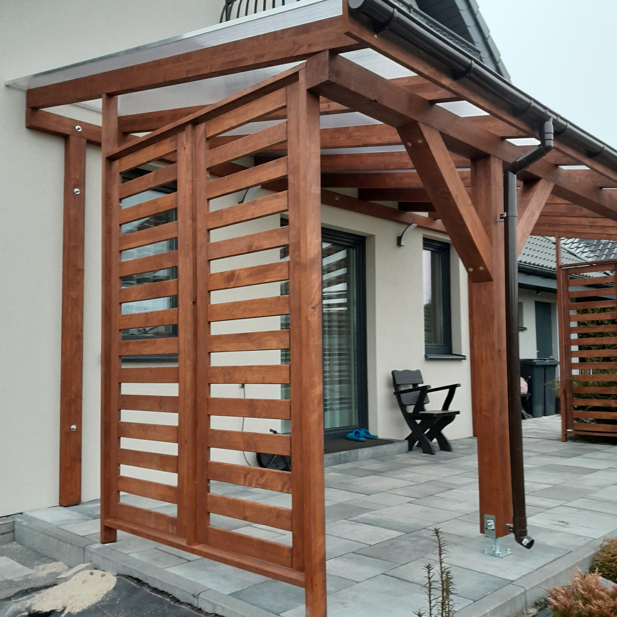 Zadaszenie tarasu z drewnianą konstrukcją i ażurowymi ściankami bocznymi, widoczne rynny, szara kostka brukowa i fragment domu.