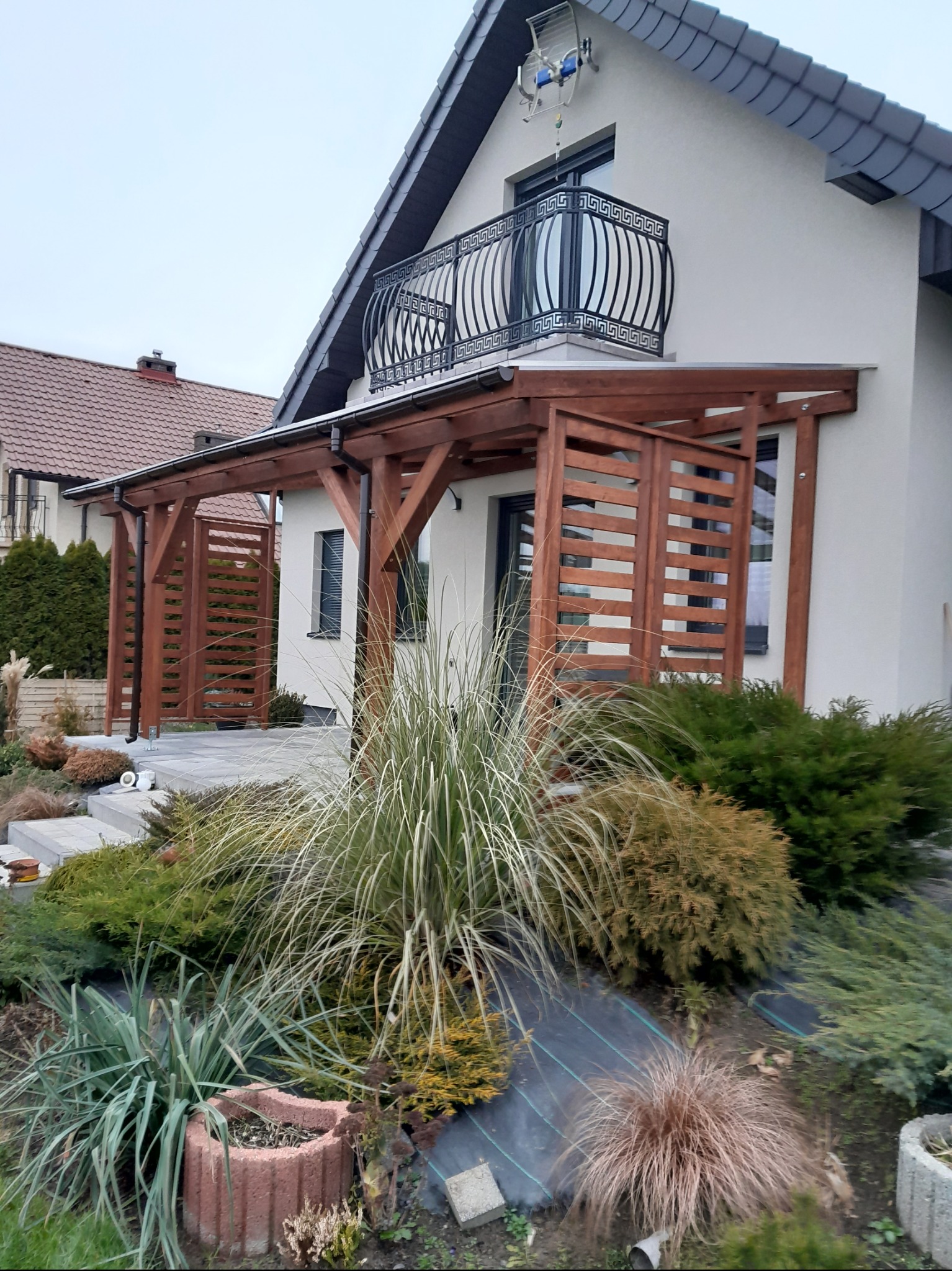 Drewniana konstrukcja z ażurowymi ściankami, przylegająca do jasnego domu z balkonem z ozdobną, metalową balustradą, otoczona zielenią i kamiennymi schodkami.