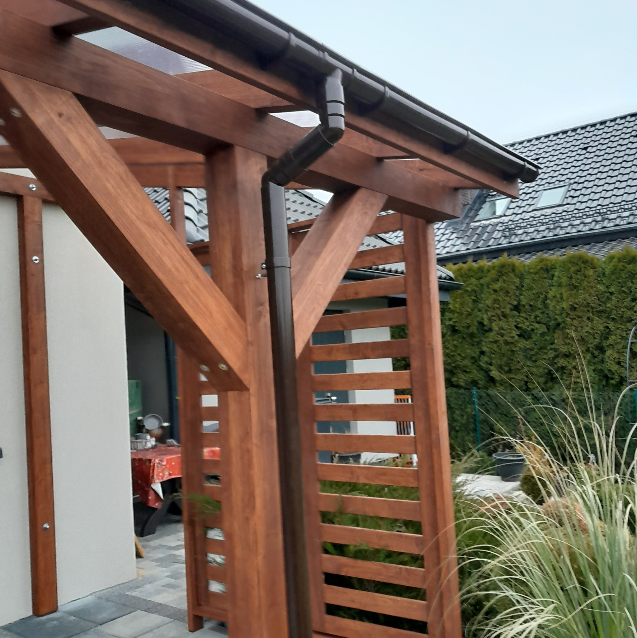 Detale konstrukcji drewnianej z brązową rynną, widoczne elementy zadaszenia i fragment domu w tle.