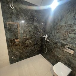 VOLTA - Wykończona kabina prysznicowa z ciemnymi, marmurkowymi płytkami, deszczownicą i podwieszaną toaletą, widoczny odpływ liniowy w podłodze.