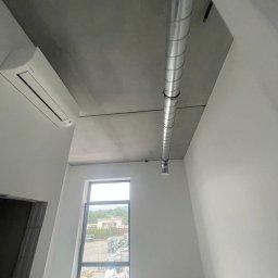 VOLTA - Wnętrze w stanie deweloperskim z widoczną instalacją wentylacyjną pod sufitem, klimatyzatorem na ścianie i oknem z widokiem na teren zewnętrzny.