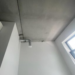 VOLTA - Szara, surowa powierzchnia sufitu z widoczną instalacją wentylacyjną i fragmentem ściany pomalowanej na biało, oświetlona naturalnym światłem z okna.
