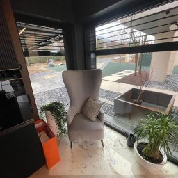 VOLTA - Jasnoszary fotel typu uszak z poduszką w rogu nowoczesnego salonu z marmurową podłogą, widok na ogród przez duże okno z żaluzjami.