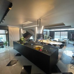 VOLTA - Przestronne wnętrze domu z nowoczesną kuchnią z wyspą z czarnego kamienia, marmurową podłogą w geometryczny wzór i schodami w tle, oświetlone ledowymi lampami.