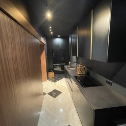 VOLTA - Wąska kuchnia w ciemnej tonacji z błyszczącymi marmurowymi płytkami podłogowymi ułożonymi w szachownicę, drewnianą ścianą i minimalistyczną zabudową kuchenną z czarnymi frontami.