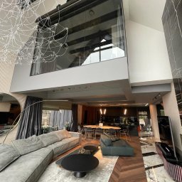 VOLTA - Przestronne wnętrze domu z antresolą, widok na salon z szarą sofą, czarnymi stolikami kawowymi i dekoracyjną lampą, parkiet w jodełkę, marmurowa posadzka w tle