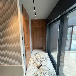 VOLTA - Nowoczesny korytarz z marmurową podłogą w odcieniach bieli i szarości, drewnianymi drzwiami i panelami ściennymi z pionowymi żłobieniami, oświetlony reflektorami na szynoprzewodzie