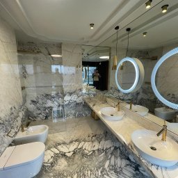 VOLTA - Elegancka łazienka wykończona marmurem z białymi umywalkami nablatowymi i okrągłymi lustrami podświetlanymi LED, widoczne elementy armatury w kolorze złotym.