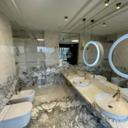 Elegancka łazienka wykończona marmurem z białymi umywalkami nablatowymi i okrągłymi lustrami podświetlanymi LED, widoczne elementy armatury w kolorze złotym.
