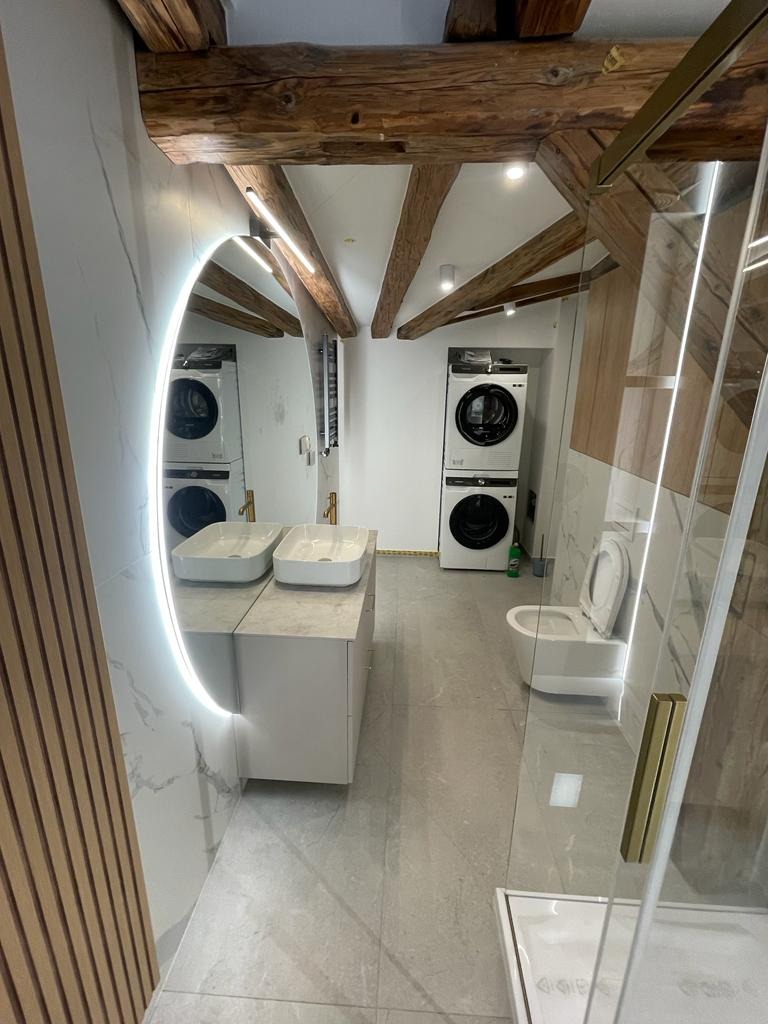 Nowoczesna łazienka z drewnianymi belkami stropowymi, podwójną umywalką nablatową, toaletą i pralko-suszarką w tle, odbicie w okrągłym lustrze z podświetleniem LED.
