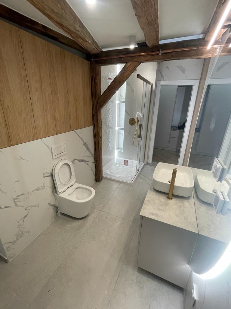 Nowoczesna łazienka z drewnianymi belkami stropowymi, marmurowymi płytkami i kabiną prysznicową ze złotymi akcentami.