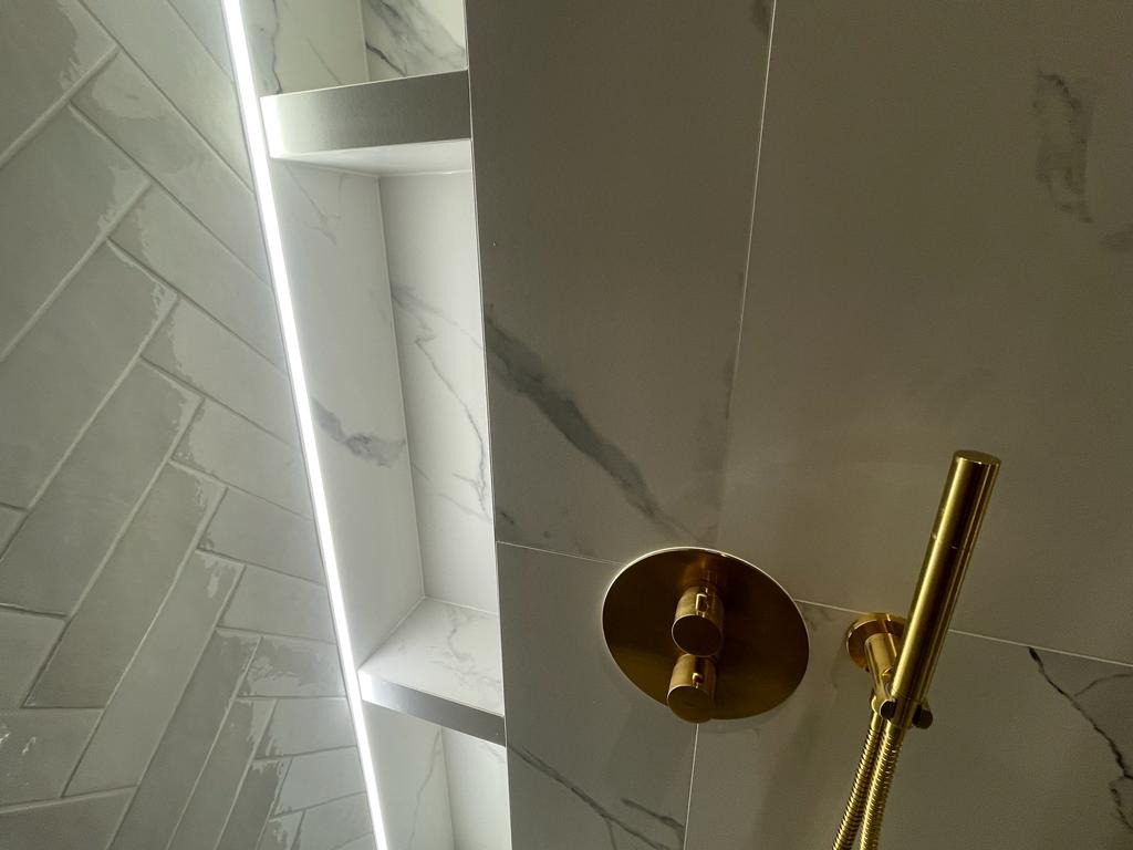 Wykończona łazienka z szarymi płytkami ułożonymi w jodełkę, wnęką z podświetleniem LED i złotą armaturą.