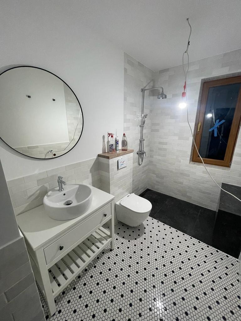 Nowoczesna łazienka z okrągłym lustrem, białą umywalką na szafce z szufladą i półkami, toaletą w zabudowie oraz kabiną prysznicową z deszczownicą; podłoga wyłożona mozaiką w biało-czarne kropki,...