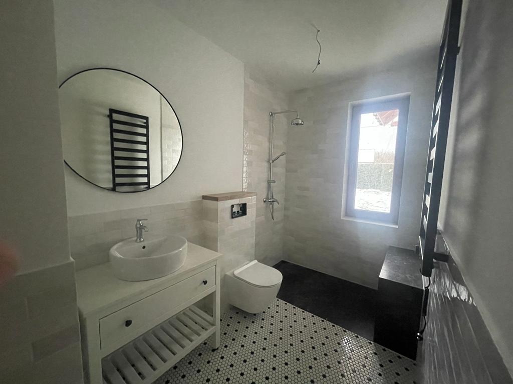 Nowoczesna łazienka z okrągłym lustrem, białą umywalką na szafce z półkami, toaletą w zabudowie, kabiną prysznicową typu walk-in i czarno-białą mozaiką na podłodze.