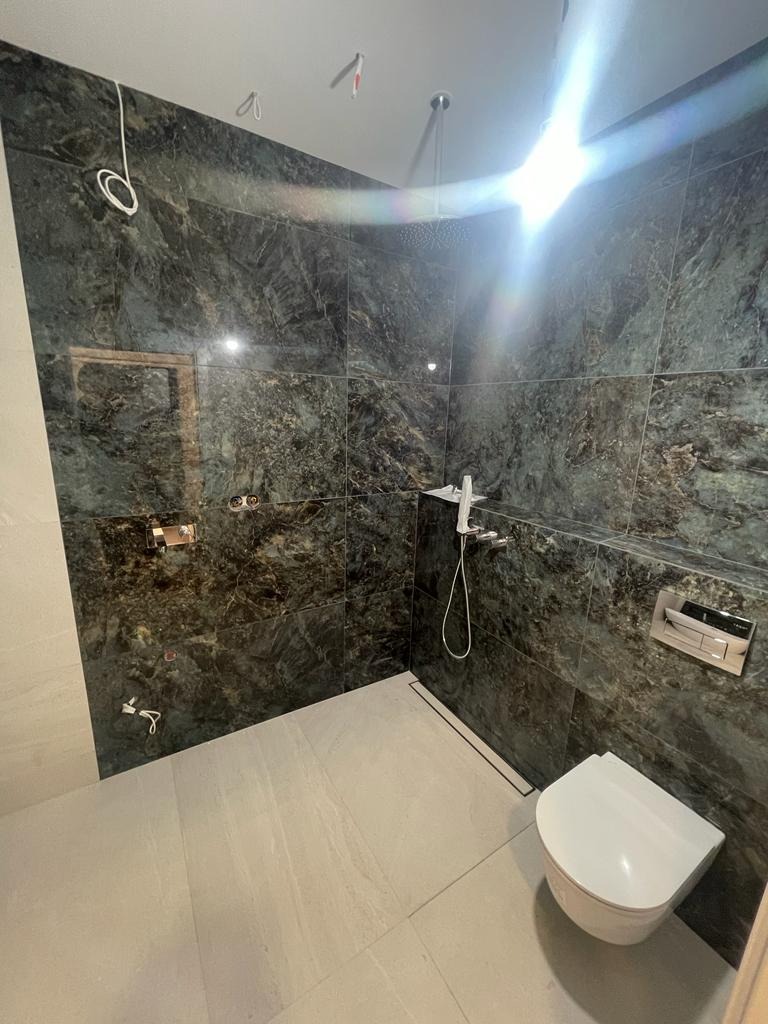 Wykończona kabina prysznicowa z ciemnymi, marmurkowymi płytkami, deszczownicą i podwieszaną toaletą, widoczny odpływ liniowy w podłodze.