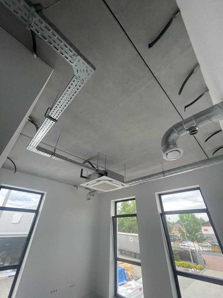 Surowy betonowy sufit z widocznymi instalacjami wentylacyjnymi, kanałami kablowymi i klimatyzacją w pustym pomieszczeniu biurowym z trzema oknami.
