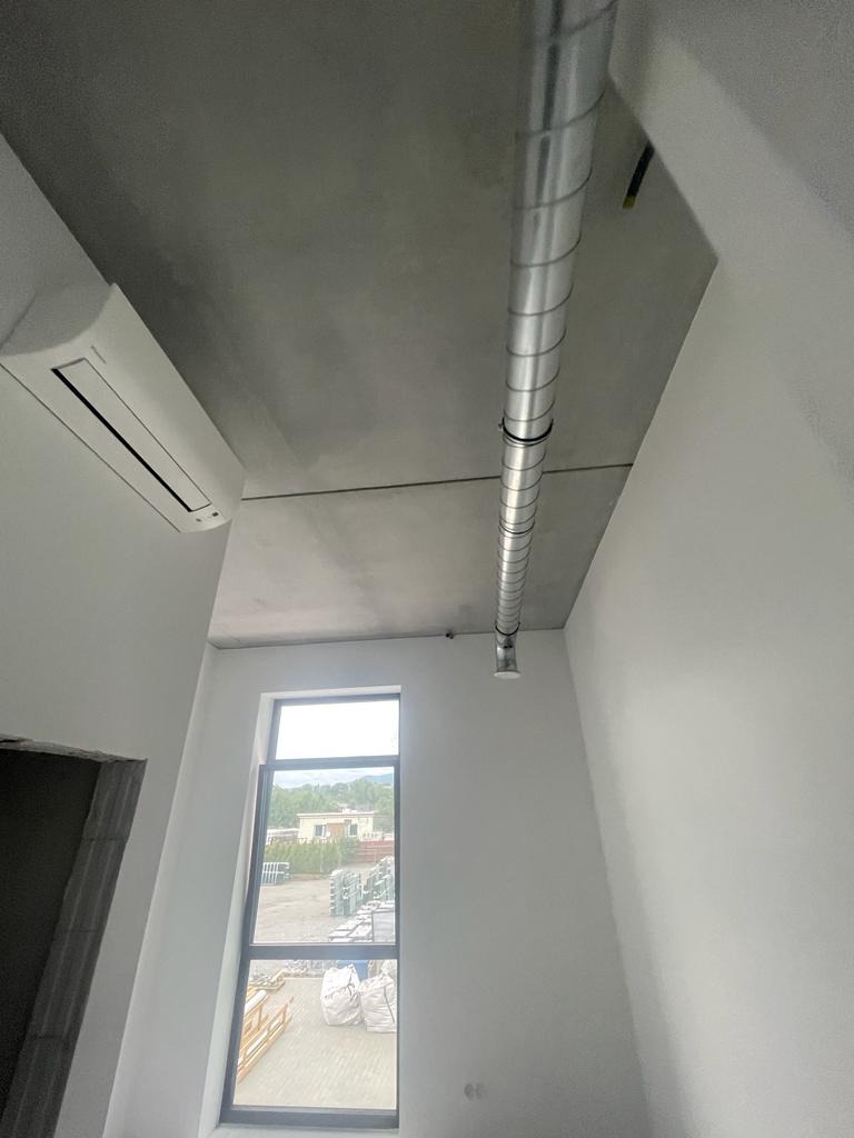 Wnętrze w stanie deweloperskim z widoczną instalacją wentylacyjną pod sufitem, klimatyzatorem na ścianie i oknem z widokiem na teren zewnętrzny.