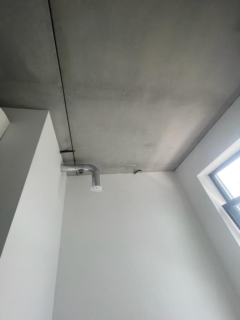 Szara, surowa powierzchnia sufitu z widoczną instalacją wentylacyjną i fragmentem ściany pomalowanej na biało, oświetlona naturalnym światłem z okna.