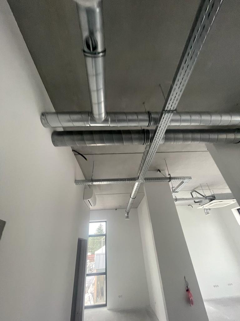 Wnętrze w stanie deweloperskim z widocznymi instalacjami wentylacyjnymi pod sufitem, klimatyzator ścienny, surowa podłoga i ściany przygotowane do wykończenia, okno z widokiem na zieleń.