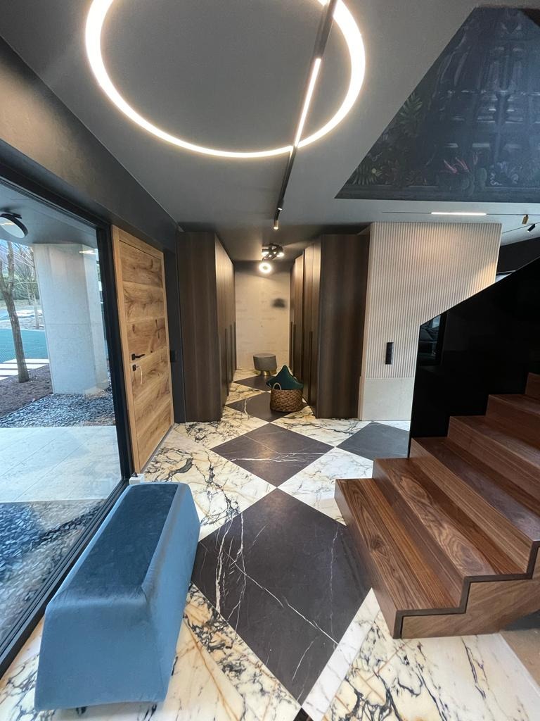 Nowoczesny hol z marmurową podłogą w szachownicę, drewnianymi schodami i szafami w zabudowie, oświetlony okrągłą lampą LED i z dekoracyjnym malowidłem na suficie.