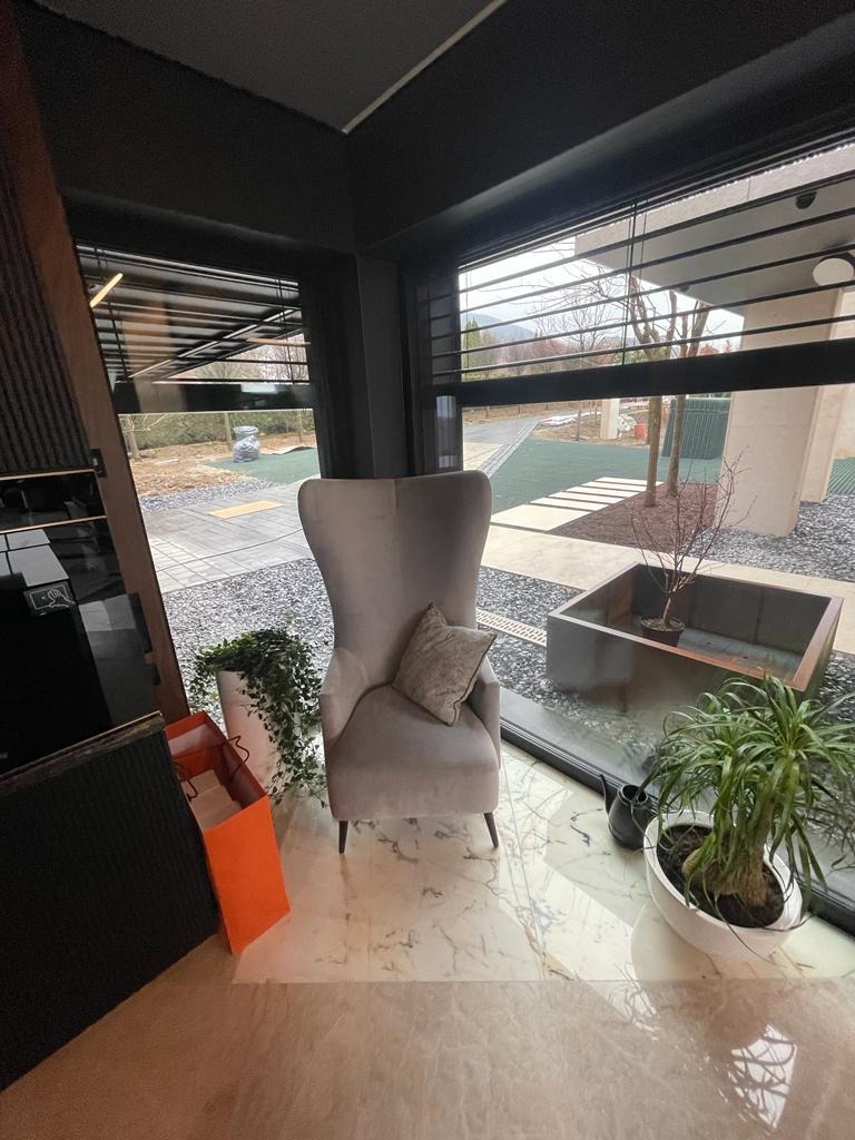 Jasnoszary fotel typu uszak z poduszką w rogu nowoczesnego salonu z marmurową podłogą, widok na ogród przez duże okno z żaluzjami.