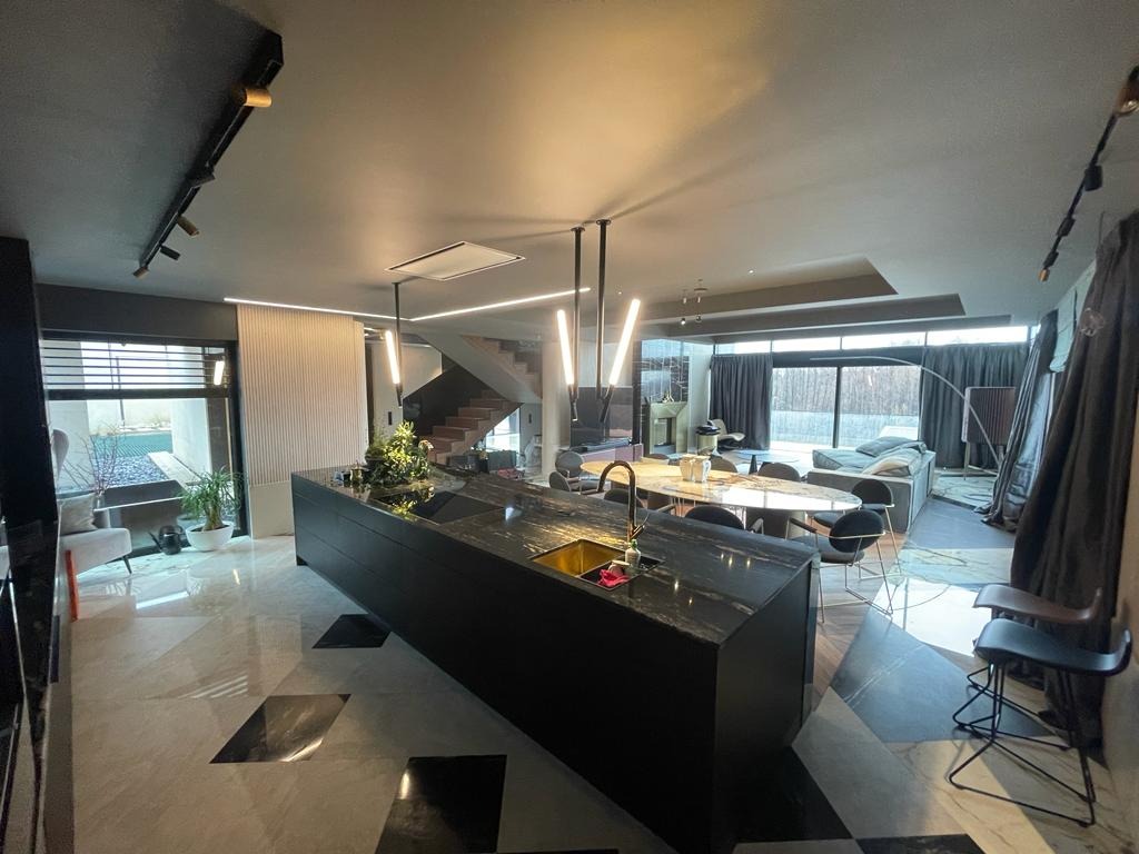 Przestronne wnętrze domu z nowoczesną kuchnią z wyspą z czarnego kamienia, marmurową podłogą w geometryczny wzór i schodami w tle, oświetlone ledowymi lampami.