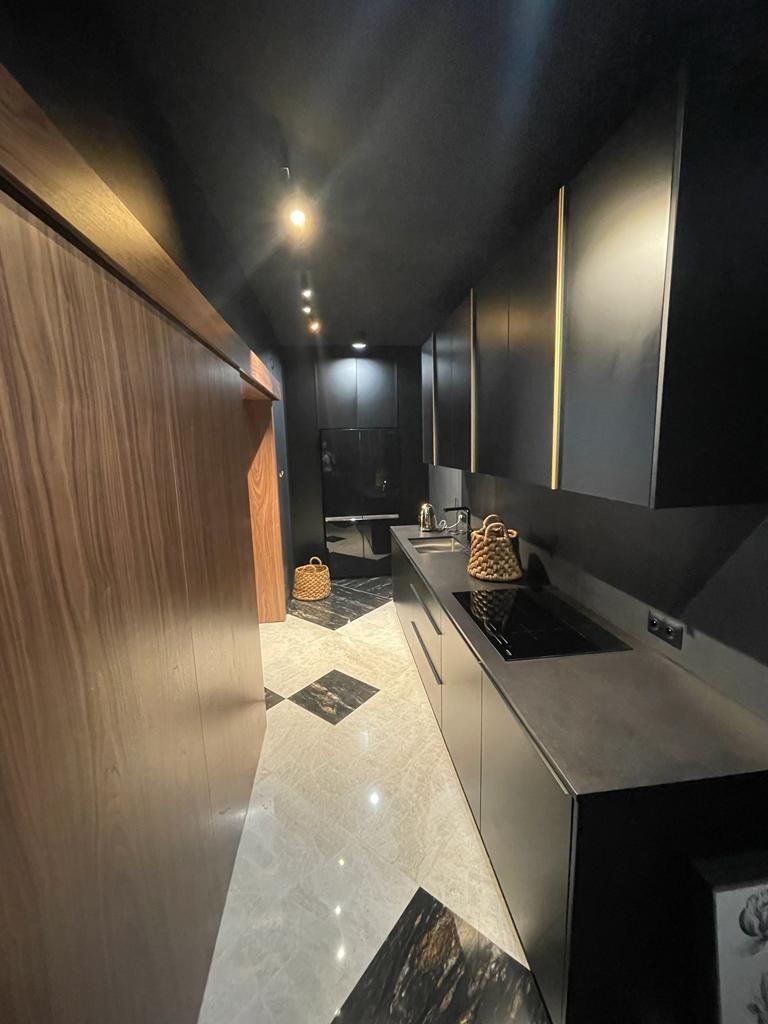 Wąska kuchnia w ciemnej tonacji z błyszczącymi marmurowymi płytkami podłogowymi ułożonymi w szachownicę, drewnianą ścianą i minimalistyczną zabudową kuchenną z czarnymi frontami.