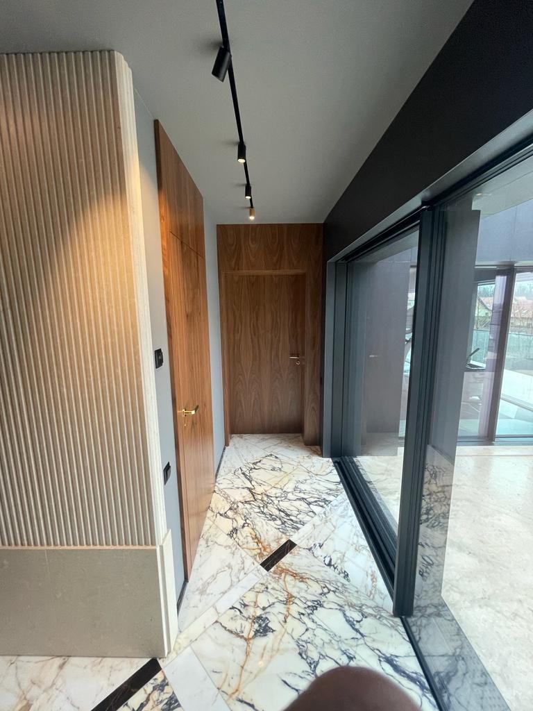 Nowoczesny korytarz z marmurową podłogą w odcieniach bieli i szarości, drewnianymi drzwiami i panelami ściennymi z pionowymi żłobieniami, oświetlony reflektorami na szynoprzewodzie