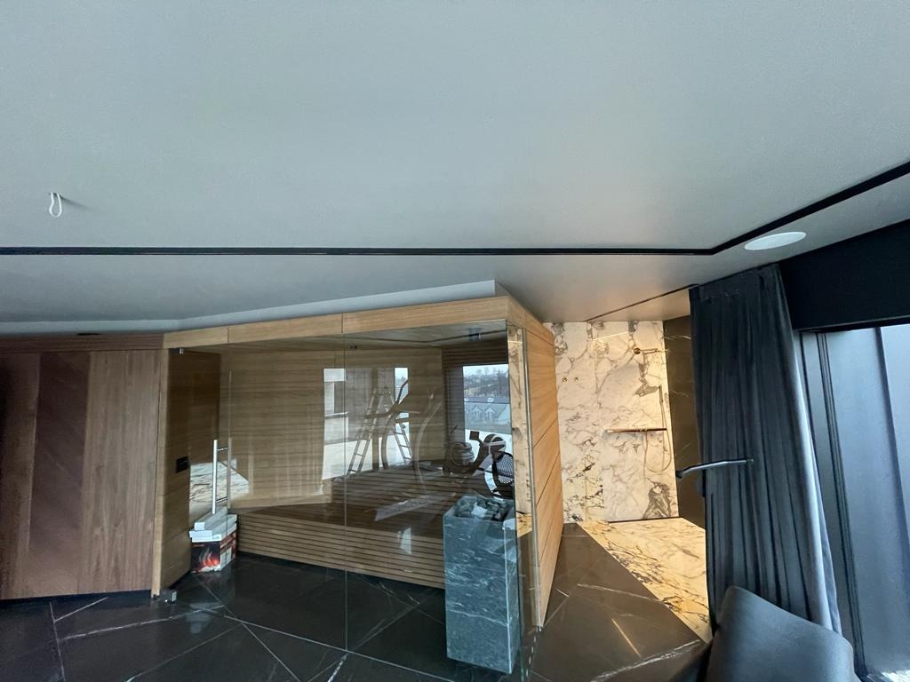 Nowoczesna łazienka z sauną: drewniana sauna za szklaną ścianą, marmurowe płytki na ścianie, czarna podłoga z połyskiem, ciemne zasłony, minimalistyczny design.