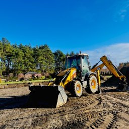 ECOBUILD-K - Firma ogólnobudowlana - Żółta koparko-ładowarka JCB na placu budowy, widoczne ślady opon na ziemi, w tle wykop i drzewa pod błękitnym niebem.