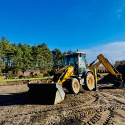 Żółta koparko-ładowarka JCB na placu budowy, widoczne ślady opon na ziemi, w tle wykop i drzewa pod błękitnym niebem.
