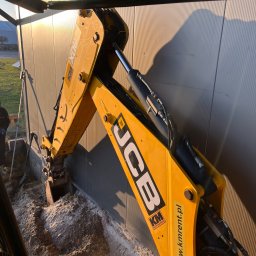 ECOBUILD-K - Firma ogólnobudowlana - Żółta koparko-ładowarka JCB wykonuje wykop wzdłuż metalowej ściany budynku, widoczna ziemia i częściowo operator maszyny.