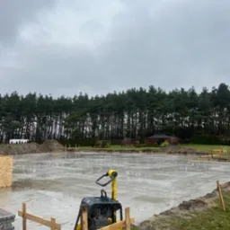 Świeżo wylana płyta betonowa pod budowę domu, widoczna zagęszczarka wibracyjna, drewniane szalunki, w tle las i pochmurne niebo.