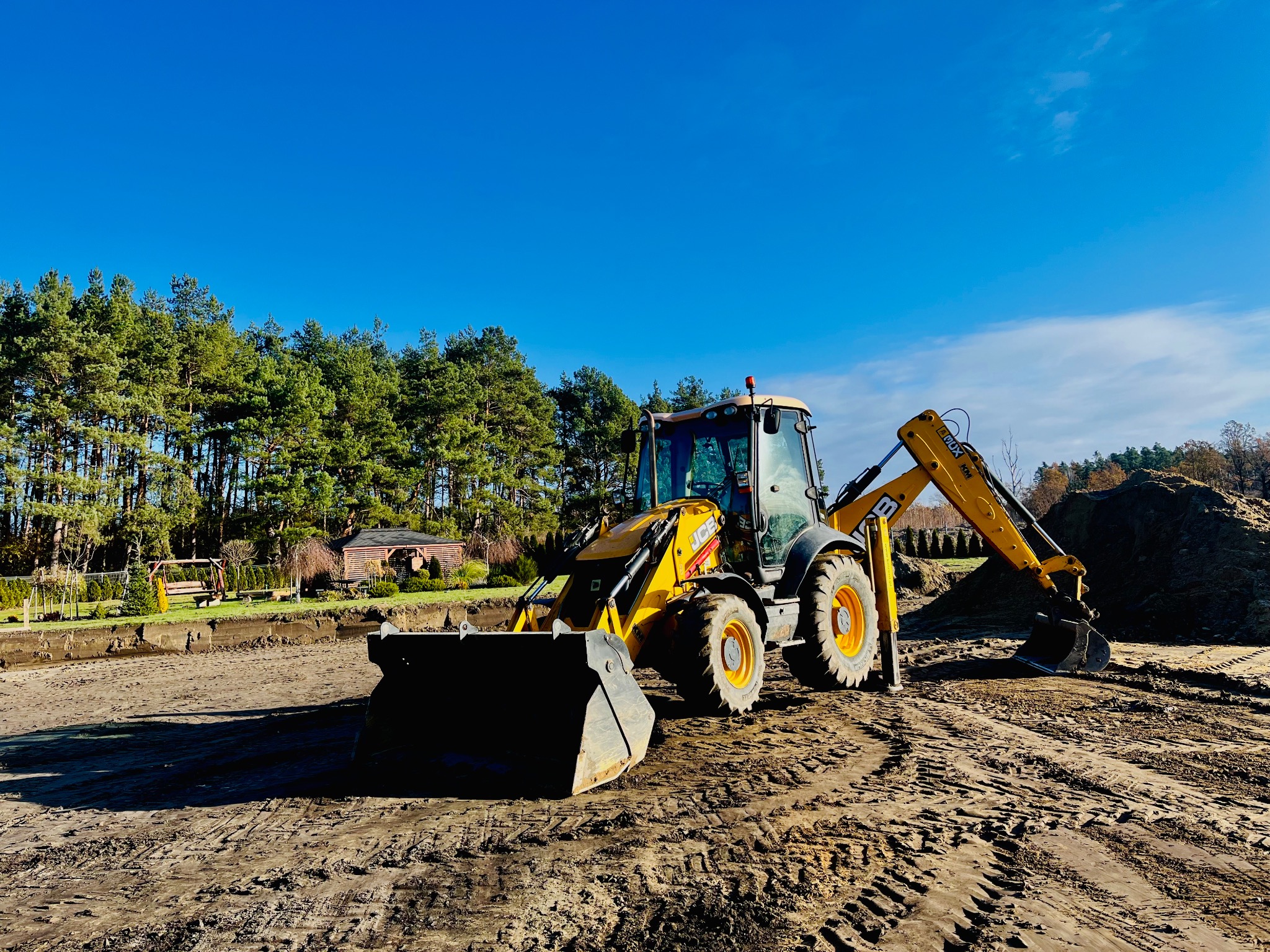 Żółta koparko-ładowarka JCB na placu budowy, widoczne ślady opon na ziemi, w tle wykop i drzewa pod błękitnym niebem.