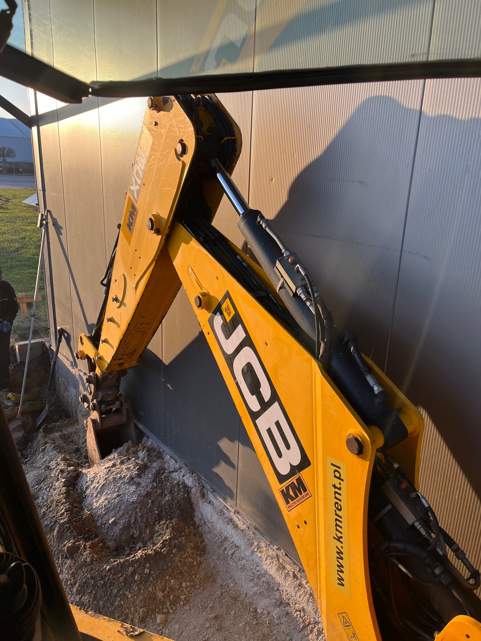 Żółta koparko-ładowarka JCB wykonuje wykop wzdłuż metalowej ściany budynku, widoczna ziemia i częściowo operator maszyny.
